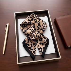 Può includere: Una sciarpa con stampa leopardata beige e nera, piegata in una confezione regalo. La sciarpa ha un bordo nero ed è realizzata in un materiale setoso. Sono visibili anche una penna dorata e un taccuino in pelle marrone.