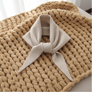 Peut inclure: Une écharpe beige nouée, posée sur une couverture en tricot épais. L'écharpe a un design simple avec des extrémités longues et pointues. La couverture est d'une couleur chaude et neutre, créant une esthétique chaleureuse et accueillante.
