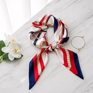 May include: A patterned silk scarf in white, red, and navy blue, knotted at the centre. A gold bracelet is placed nearby. The scarf features a repeating design with gold accents. It rests on a white marble surface.