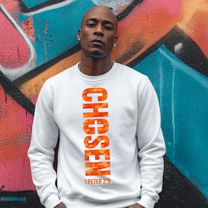 Può includere: Felpa bianca con la parola "CHOSEN" in lettere verticali arancioni mimetiche. Sotto c'è il testo "1 PETER 2:9". Lo sfondo è un colorato muro di graffiti.