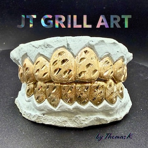 May include: Gold-colored dental grills with a textured, openwork design, displayed on a light gray mold. The text "JT GRILL ART" is at the top, with "by Thomas K." at the bottom. The grills are a dental fashion accessory.