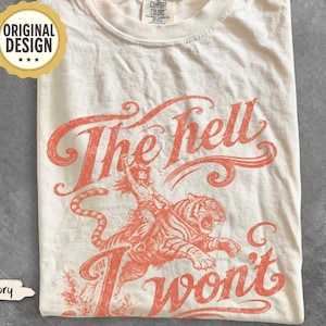 May include: An ivory short-sleeve t-shirt featuring a coral-colored graphic print of a person riding a tiger. The text "The hell I won't" is printed in a stylized script font above and below the illustration. A Comfort Colors label is at the neck.