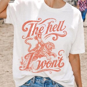 May include: A white short-sleeve t-shirt featuring a red graphic print of a tiger and the text The hell I won't. The shirt is paired with blue denim shorts. The graphic design includes stylized lettering and a tiger illustration on the front.