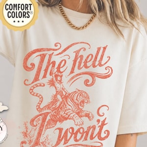 May include: An ivory short-sleeve t-shirt featuring a distressed red graphic of a person riding a tiger. The text reads The hell I won't in a stylized script font. A circular Comfort Colors logo is visible in the upper left corner of the image.