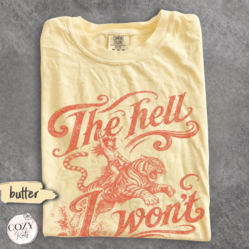 May include: A butter yellow short-sleeve t-shirt featuring a distressed red graphic of a person riding a tiger. The text reads The hell I won't in a stylised script font. The shirt has a Comfort Colors brand label at the inner neckline.