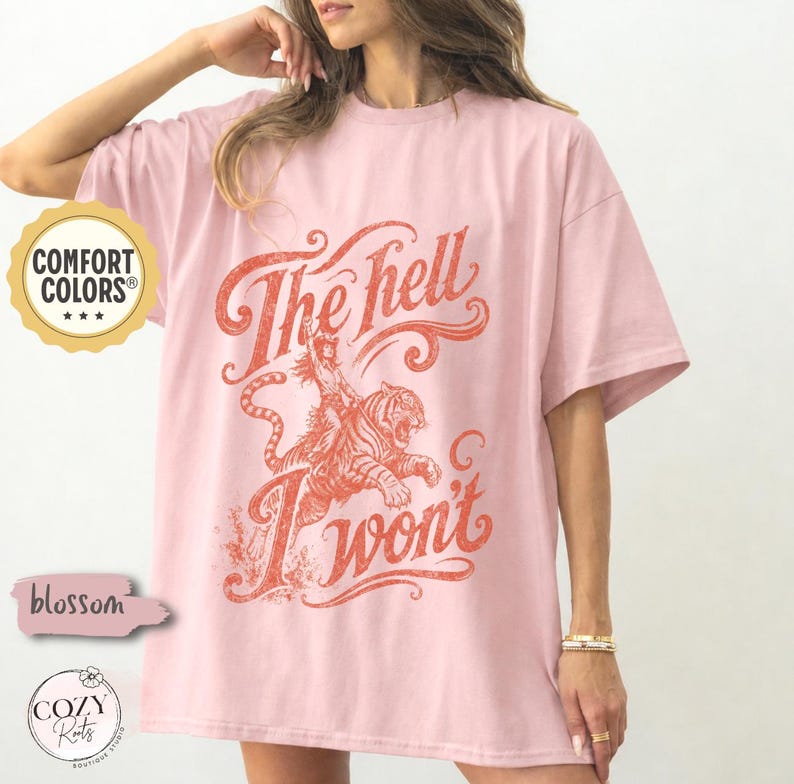 May include: A light pink short-sleeve t-shirt featuring a distressed orange graphic of a person riding a tiger. The text reads The hell I won't in a stylised script font. The shirt has a crew neckline and a loose, relaxed fit throughout.