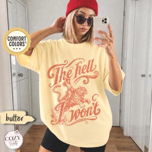 May include: A butter yellow short-sleeve t-shirt featuring a red graphic print of a person riding a tiger with the text "The hell I won't" in a stylised font. The shirt has a crew neckline and a loose, relaxed fit with a vintage-inspired design.