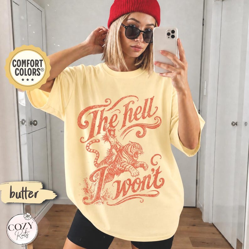 May include: A butter yellow short-sleeve t-shirt featuring a red graphic print of a person riding a tiger with the text "The hell I won't" in a stylised font. The shirt has a crew neckline and a loose, relaxed fit with a vintage-inspired design.