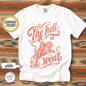 May include: A white short-sleeve t-shirt featuring a red graphic print of a person riding a tiger. The text "The hell I won't" is written in a stylised script font above and below the illustration. The shirt has a crew neckline and a Comfort Colors label.