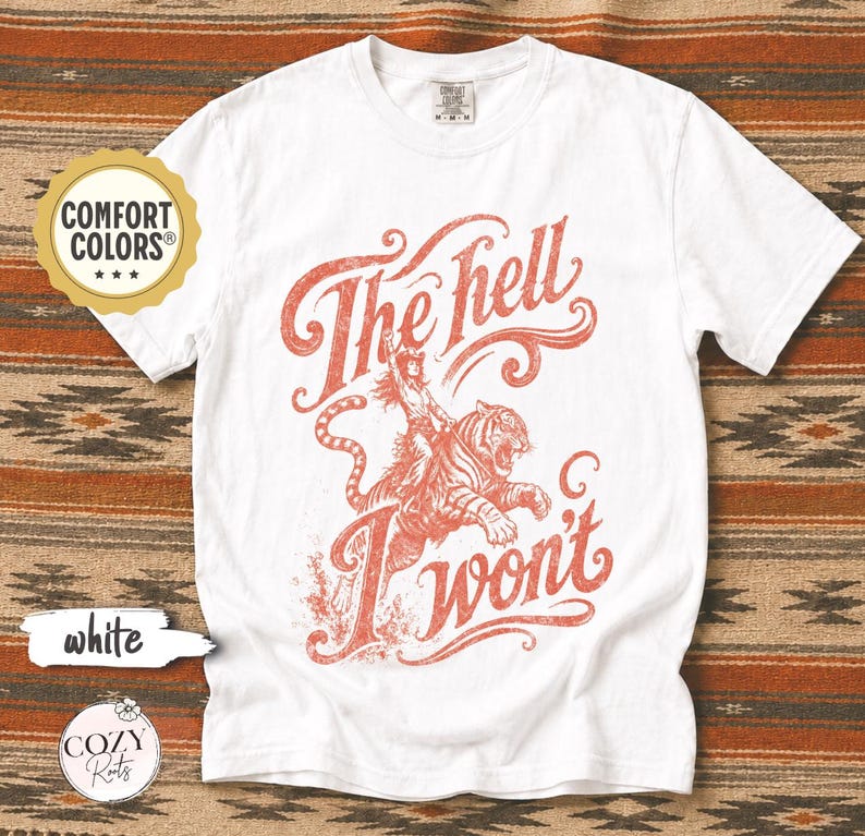 May include: A white short-sleeve t-shirt featuring a red graphic print of a person riding a tiger. The text "The hell I won't" is written in a stylised script font above and below the illustration. The shirt has a crew neckline and a Comfort Colors label.