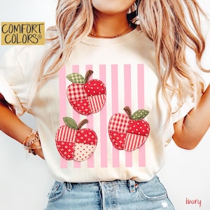 May include: An ivory t-shirt featuring a graphic of three apples made of patchwork fabric patterns in shades of red, white, and green. The apples are layered over a background of vertical light pink stripes. The text COMFORT COLORS appears in the corner.