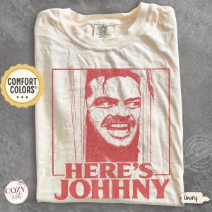 May include: An ivory t-shirt featuring a red graphic print of a man's face peering through a broken door. The text "HERE'S JOHNNY" is printed in red below the image. The shirt has a Comfort Colors brand label at the inner neckline.