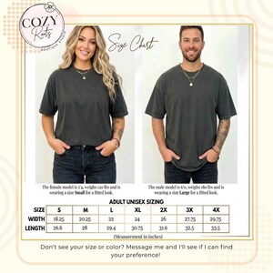 May include: A size chart for a charcoal grey unisex t-shirt featuring two models wearing the shirt. The chart displays adult unisex sizing in inches, ranging from size S to 4X, with specific width and length measurements for each size provided.