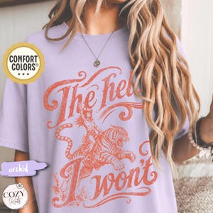 May include: A light purple short-sleeve t-shirt featuring a graphic print in a faded orange hue. The design depicts a person riding a tiger with the text "The hell I won't" in a stylized script font. A Comfort Colors brand logo is visible.