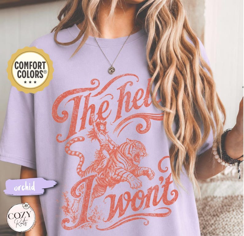 May include: A light purple short-sleeve t-shirt featuring a graphic print in a faded orange hue. The design depicts a person riding a tiger with the text "The hell I won't" in a stylized script font. A Comfort Colors brand logo is visible.