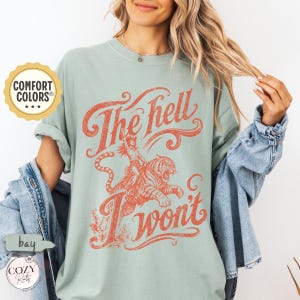 May include: A sage green short-sleeve t-shirt featuring a distressed orange graphic of a person riding a tiger. The text reads "The hell I won't" in a stylised, vintage-inspired font. The shirt has a crew neckline and a relaxed, loose fit.