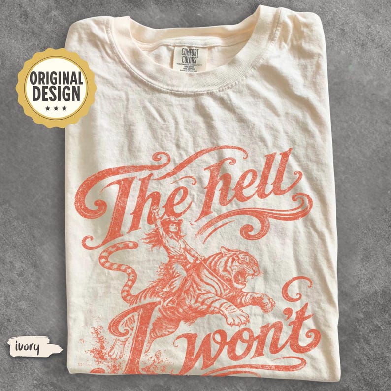 May include: An ivory short-sleeve t-shirt featuring a coral-colored graphic print of a person riding a tiger. The text "The hell I won't" is printed in a stylized script font above and below the illustration. A Comfort Colors label is at the neck.