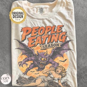 May include: A cream-colored t-shirt featuring a graphic print with the text "PEOPLE EATING SEASON" in orange. The design depicts a purple, one-eyed winged monster with horns looming over a person in a white dress against a large orange moon.