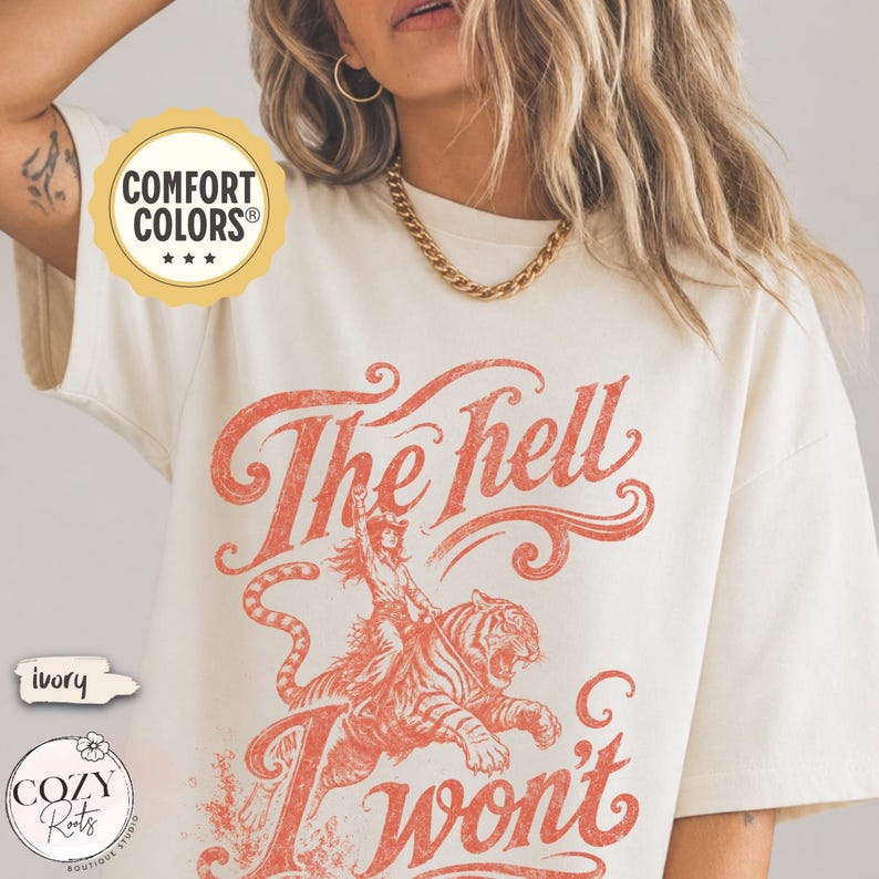 May include: An ivory short-sleeve t-shirt featuring a distressed red graphic of a person riding a tiger. The text reads The hell I won't in a stylized script font. A circular Comfort Colors logo is visible in the upper left corner of the image.