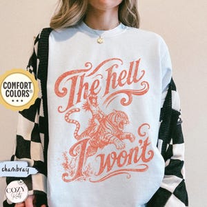 May include: A light blue crewneck sweatshirt featuring a graphic print in a faded red hue. The design depicts a person riding a tiger with the text "The hell I won't" in a stylised script font. The garment has long sleeves and a ribbed neckline.