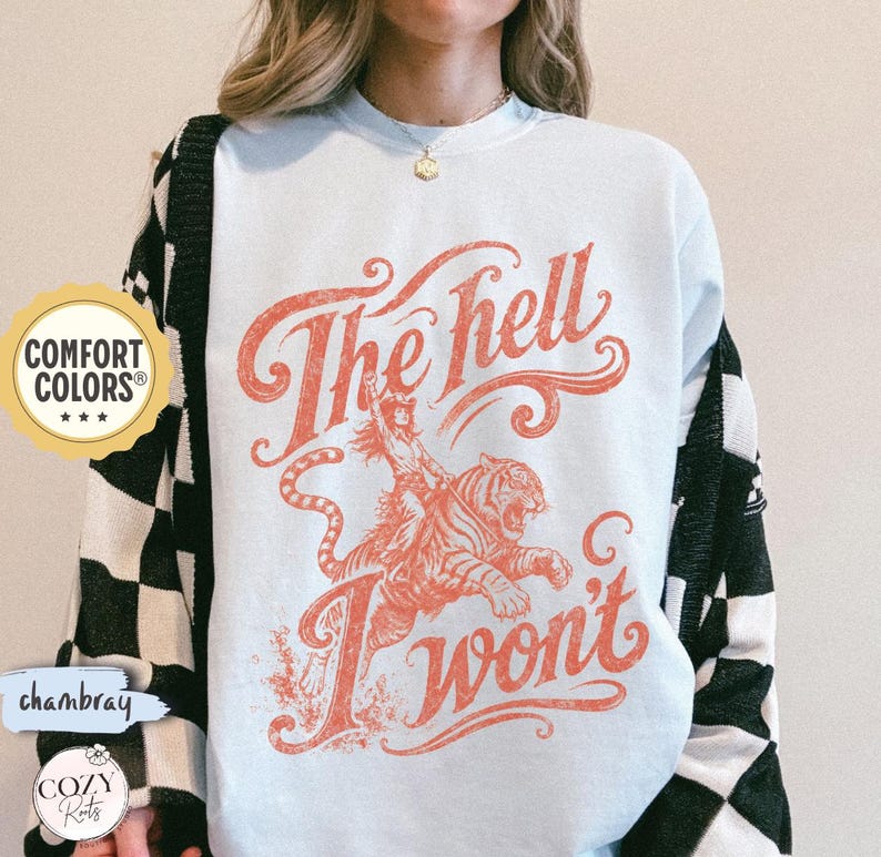May include: A light blue crewneck sweatshirt featuring a graphic print in a faded red hue. The design depicts a person riding a tiger with the text "The hell I won't" in a stylised script font. The garment has long sleeves and a ribbed neckline.