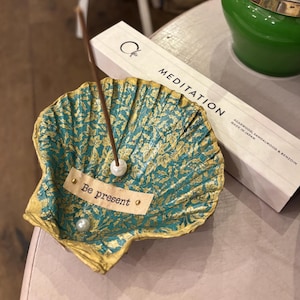 May include: A decorative seashell incense holder painted in teal and gold with a leaf pattern. It features a small white paper strip with the text Be present, two small white pearl-like beads, and a thin brown incense stick standing upright.