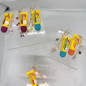 Könnte beinhalten: Schlüsselanhänger mit Miniatur-Lippenbalsam-Tuben in Gelb und farbigen Kappen. Jeder Schlüsselanhänger hat einen goldenen Verschluss und dekorative Charms, darunter Blumen, Kirschen und Blätter. Die Tuben haben Textetiketten in Rot und Gelb.