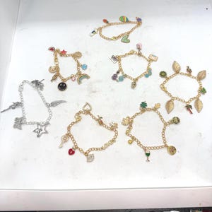 May include: A selection of charm bracelets with diverse pendants, such as a rainbow, a key, a heart, and a four-leaf clover. The bracelets are gold and silver-toned, with colourful enamel details. Charms include a smiley face, a star, and a musical note.