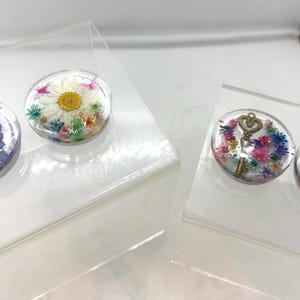 May include: Clear resin plugs with dried flowers and decorative elements. One pair features purple flowers and butterflies, another with a daisy. The other two have a key and a cross. These are displayed on clear acrylic stands.