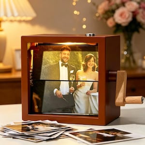 May include: A brown wooden photo cube with a hand crank, showing a wedding photograph. The cube is illuminated by an internal light, creating a warm ambience. A pile of photos is in front of the cube, indicating a collection of memories.