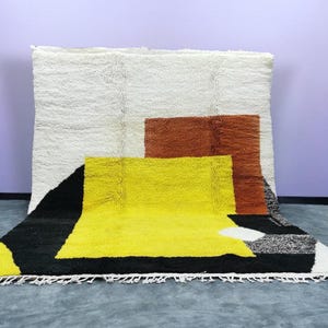 May include: A large, rectangular rug with a geometric design. The rug features a white background with a brown square and a yellow rectangle. A black triangle and white fringe complete the design.