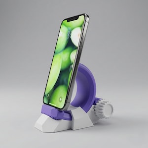 Puede incluir: Un smartphone con un fondo de pantalla de manzanas verdes se muestra en un soporte para teléfono morado y blanco. El soporte tiene un soporte circular morado, una base blanca y una perilla de ajuste en forma de engranaje. El teléfono está ligeramente inclinado.