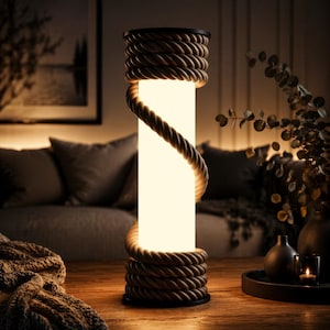 Puede incluir: Lámpara cilíndrica con un interior cálido y brillante. La lámpara está envuelta con una cuerda gruesa y texturizada, creando una estética náutica o rústica. La lámpara se asienta sobre una base oscura y tiene una parte superior oscura. La lámpara está sobre una mesa de madera.