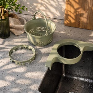 Puede incluir: Un juego de colador de fregadero de cocina verde oliva. El juego incluye un colador pequeño con asa, un anillo de drenaje circular y un colador montado en el fregadero. También se ve un dispensador de jabón de vidrio verde oscuro con bomba dorada.