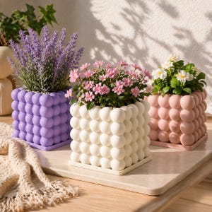 May include: Three square planters with a bubble-like texture, each filled with plants. One planter is lavender, one is off-white, and the third is a soft pink. The plants include lavender, pink flowers, and white flowers.