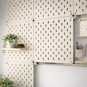 Puede incluir: Sistema de pared de panel perforado de color crema con múltiples paneles, cada uno con agujeros espaciados uniformemente. Una pequeña estantería sostiene una planta en maceta y un artículo de cuero enrollado. Accesorios adicionales: una pequeña cuadrícula enmarcada y un portalápices de tela.