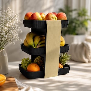 Puede incluir: Un frutero negro de tres niveles con textura acanalada vertical y una columna de soporte central ancha de color beige claro. Los estantes están llenos de varias frutas frescas, incluyendo manzanas rojas, plátanos amarillos, uvas verdes y una piña.