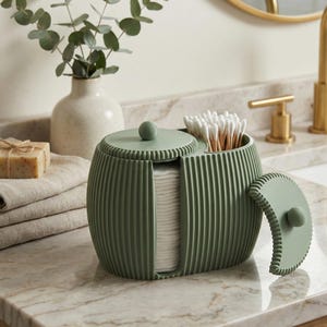 May include: A sage green bathroom organizer with a ribbed design. The organizer has a section for cotton pads and another for cotton swabs. The lid is open, revealing the contents. The organizer sits on a marble countertop.