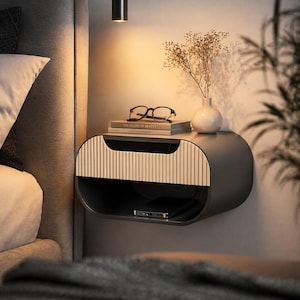 Puede incluir: Una mesita de noche moderna, montada en la pared, en un acabado gris oscuro. El frente del cajón tiene un diseño vertical y acanalado en color beige claro. Un libro, gafas, un pequeño jarrón con flores secas y un smartphone descansan encima.
