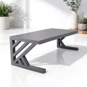 Puede incluir: Un soporte para monitor gris de estilo moderno con una parte superior rectangular y patas de soporte geométricas. El soporte mide aproximadamente 38 cm de largo, 20 cm de ancho y 10 cm de alto. Está hecho de un material mate sólido.