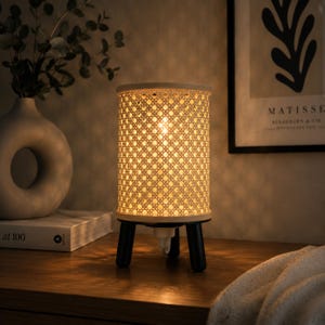 May include: A cylindrical table lamp featuring a white woven cane shade with a diamond pattern. The lamp is supported by three short, black, cylindrical legs and emits a warm, soft glow from the interior light source, casting shadows.