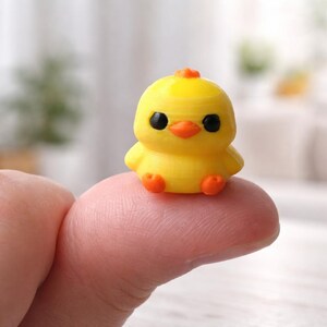 May include: A small, yellow chick figurine with black eyes and an orange beak and feet. The chick has a small orange crest on its head. The figurine is sitting on a finger, showcasing its small size. The chick is a cute, miniature decorative item.