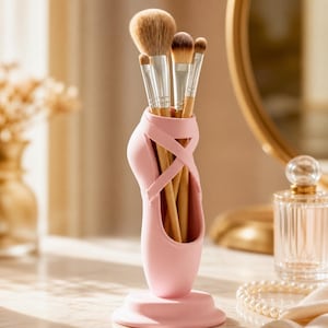 Puede incluir: Un soporte rosa en forma de zapatilla de ballet para brochas de maquillaje con varias brochas dentro. El soporte está sobre una mesa con un espejo y un frasco de perfume al fondo. Las brochas tienen mangos plateados y cerdas marrones.