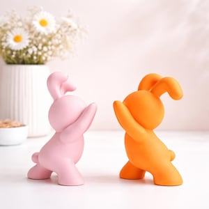 May include: Two colorful bunny figurines. One is light pink, the other is bright orange. Both are in a playful pose with one arm raised. The figurines are on a white surface, with a vase of flowers in the background.