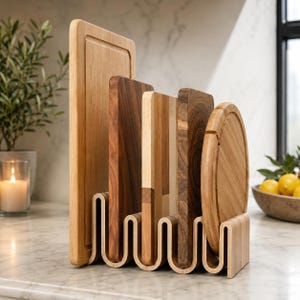 Puede incluir: Un juego de tablas de cortar de madera con una variedad de formas y vetas. Las tablas se muestran en un estante de madera ondulado sobre una encimera de mármol. Incluye tablas rectangulares y redondas.