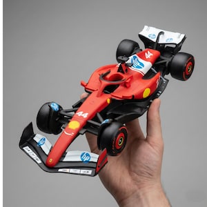 May include: A red Formula 1 race car model with black wheels and accents. The car features the number 44, sponsor logos, and a white rear wing. The model is held against a gray background.