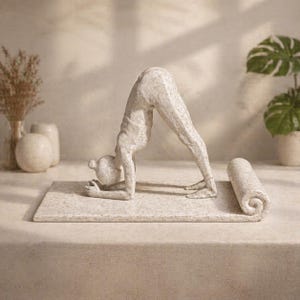 May include: A stone-colored sculpture of a person in the downward-facing dog yoga pose. The figure is on a rectangular mat, with a rolled-up yoga mat nearby. The sculpture has a textured surface and is set against a neutral background.