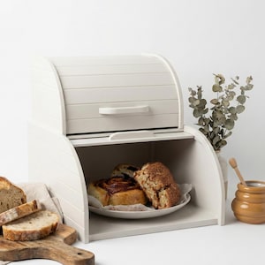 May include: A white wooden bread box with a roll-top lid and a handle. The open compartment displays a plate with a cinnamon roll and a slice of bread. A wooden cutting board with bread slices is on the left.