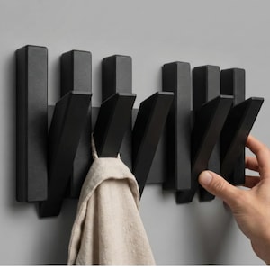 May include: A black wall-mounted coat rack with multiple folding hooks. The hooks are in the down position, and one holds a beige towel. The rack is mounted on a gray wall. A hand is reaching for one of the hooks.