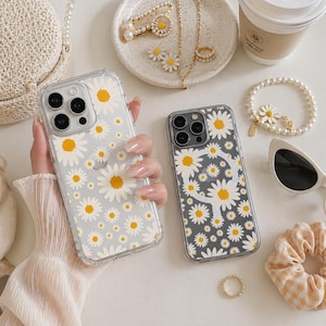 May include: Two clear phone cases with daisy flower patterns. One case is held in a hand. The daisies have white petals and yellow centers. Other accessories include jewelry, sunglasses, and a coffee cup.